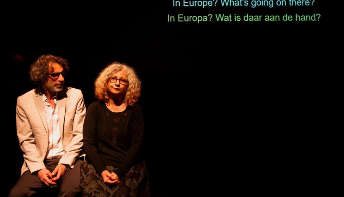 Zwei Personen sitzen auf einer Bank und sprechen miteinander. An der Wand im Hintergrund ist Schrift Projiziert.