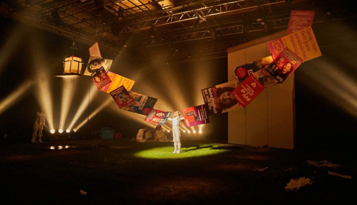 Performer_in einem Bühnenraum mit Papierflügeln und Scheinwerfern