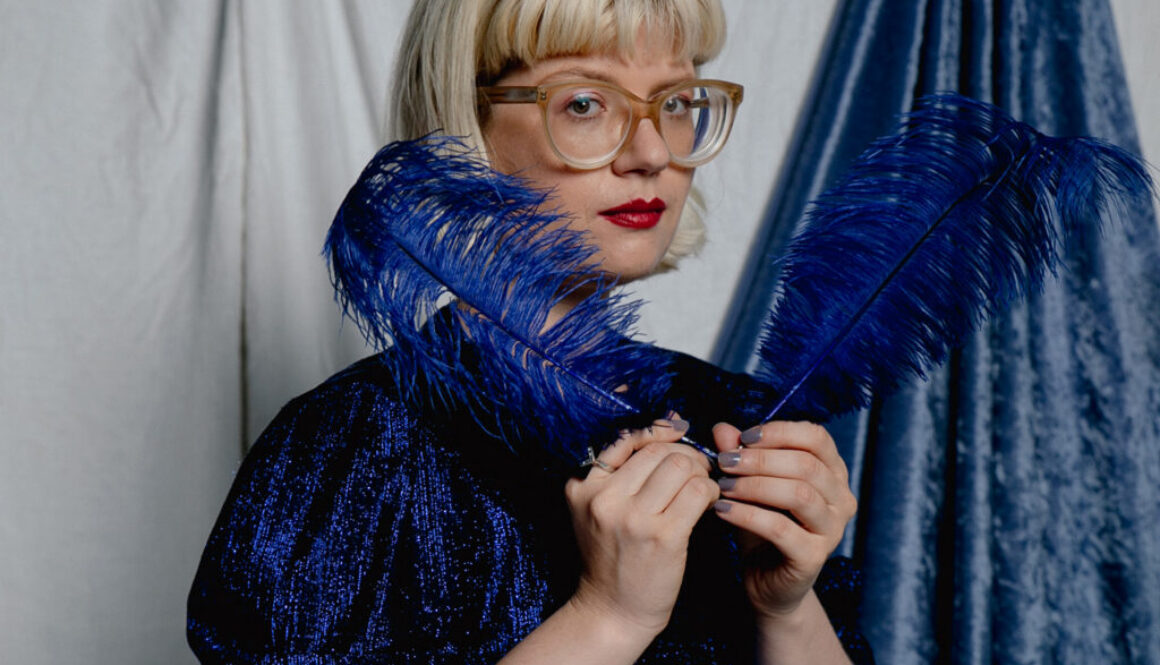 Eine Person mit Blonden Haaren und 2 blauen Federn in der Hand.